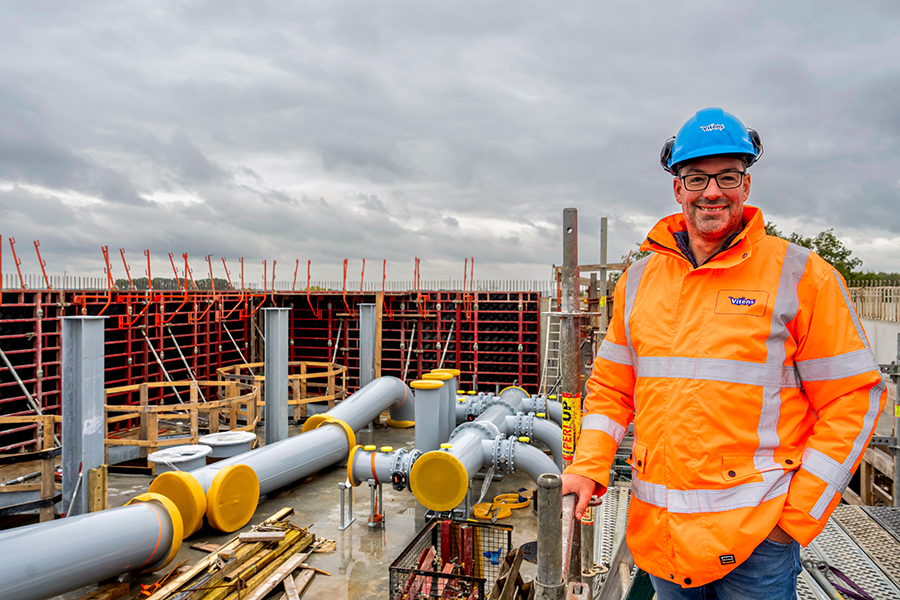 Man staat in werkjas met helm op grote bouwplaats met enorme leidingen