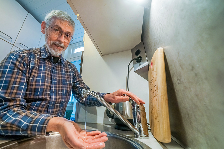 Man wast handen onder kleine straal water en kijkt lachend in de camera.