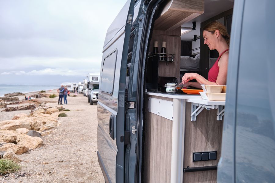 Vrouw gebruikt water in het keukentje van haar caravan. Ze kijkt uit op een strand.