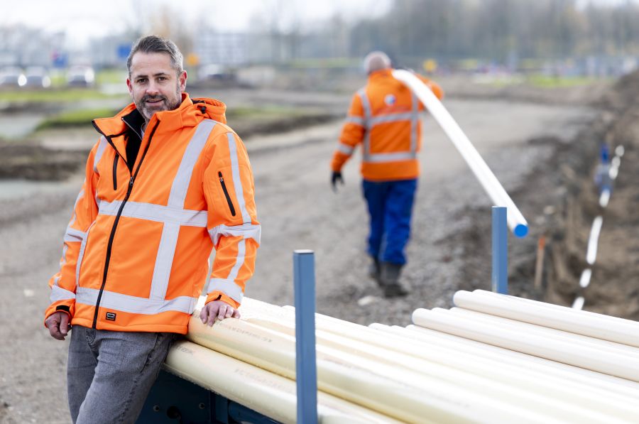 Man leunt tegen stapel drinkwaterleidingen. Op de achtergrond loopt een man weg met een drinkwaterleiding over zijn schouder.